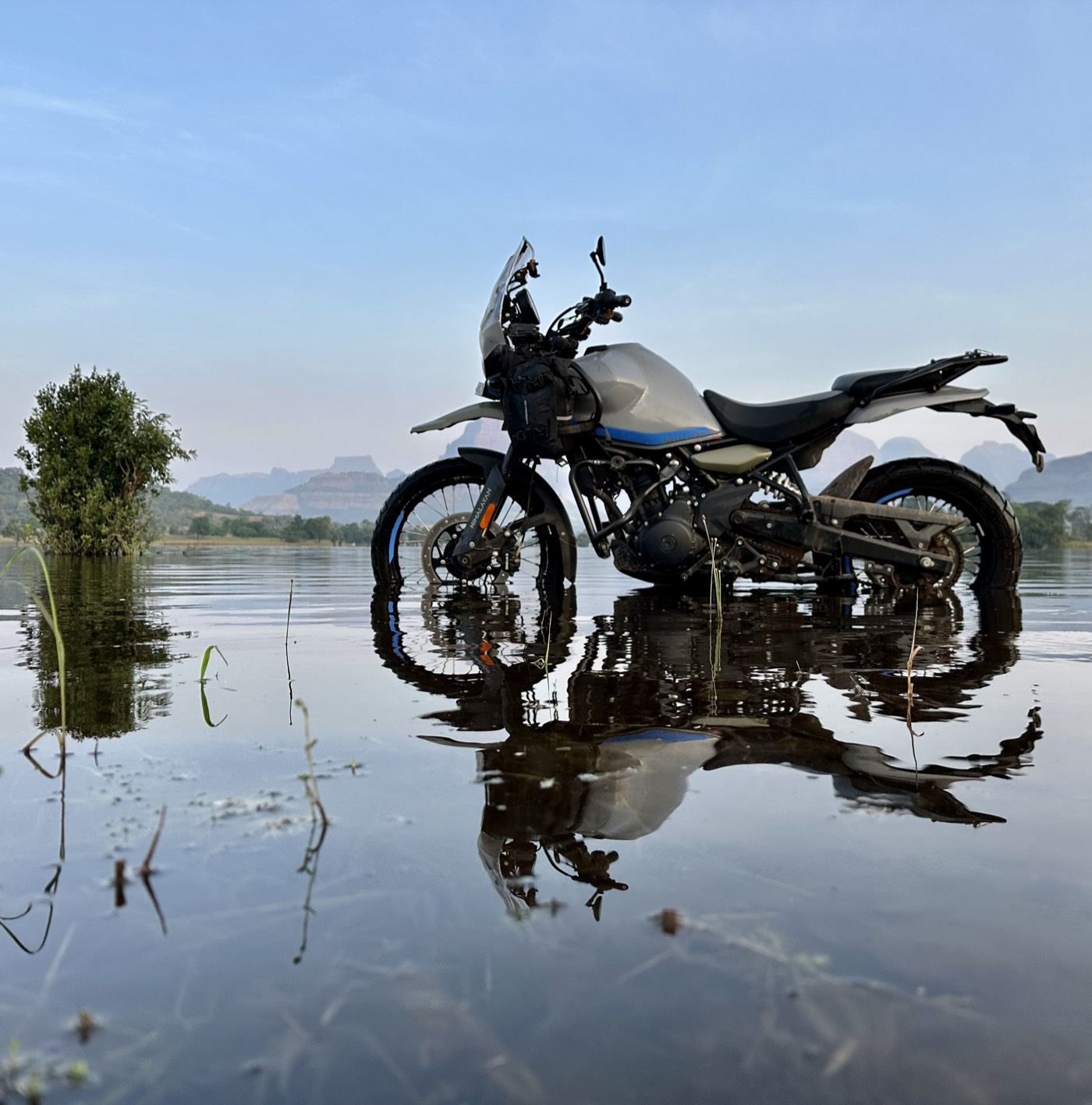 A motorcycle parked and ready, reflected in floodwater at Bhandardara with the Sahyadri mountains behind